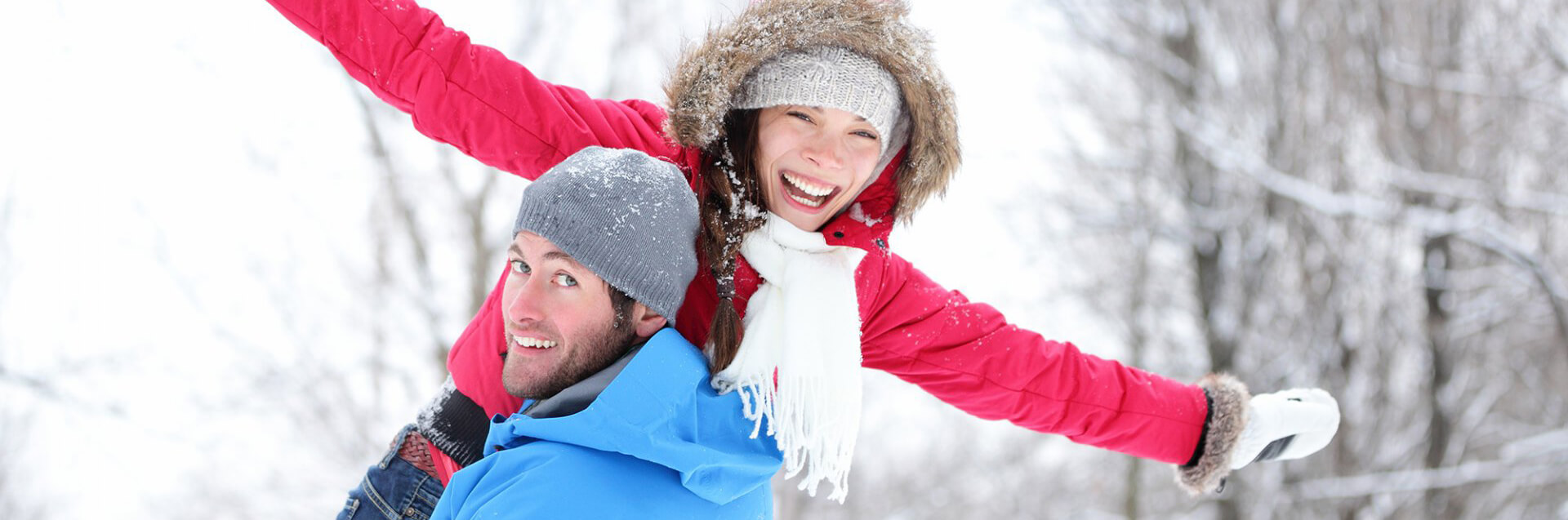 Man carrying smiling woman wearing snow clothes with snowy mountain scene in background.