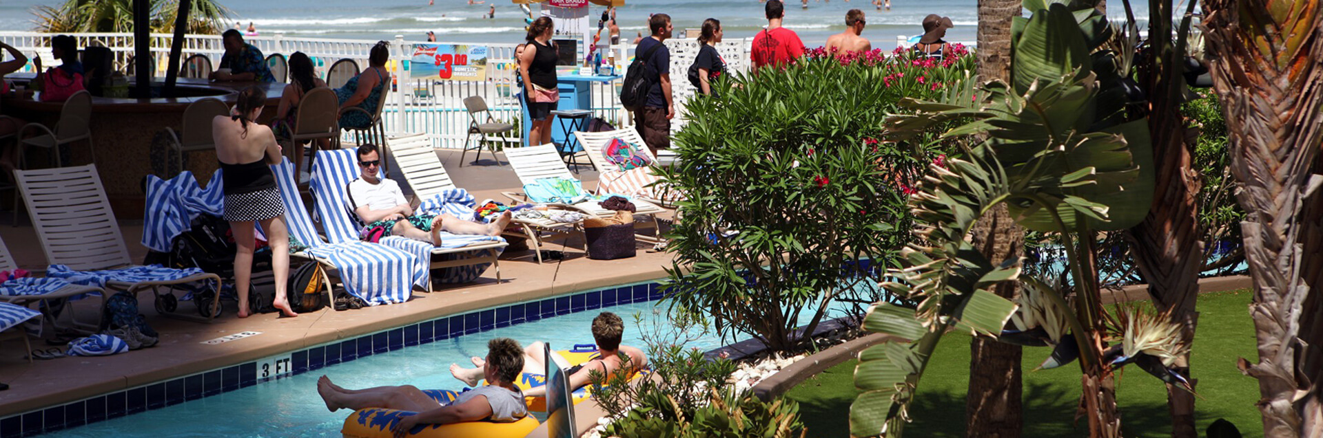 Tourists gathered around a pool at a hotel with the ocean in the background.